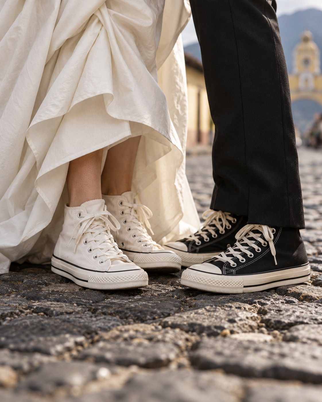 Una novia y un novio con tenis blancos y negros sobre una calle empedrada de Antigua Guatemala, con el Arco de Santa Catalina al fondo.