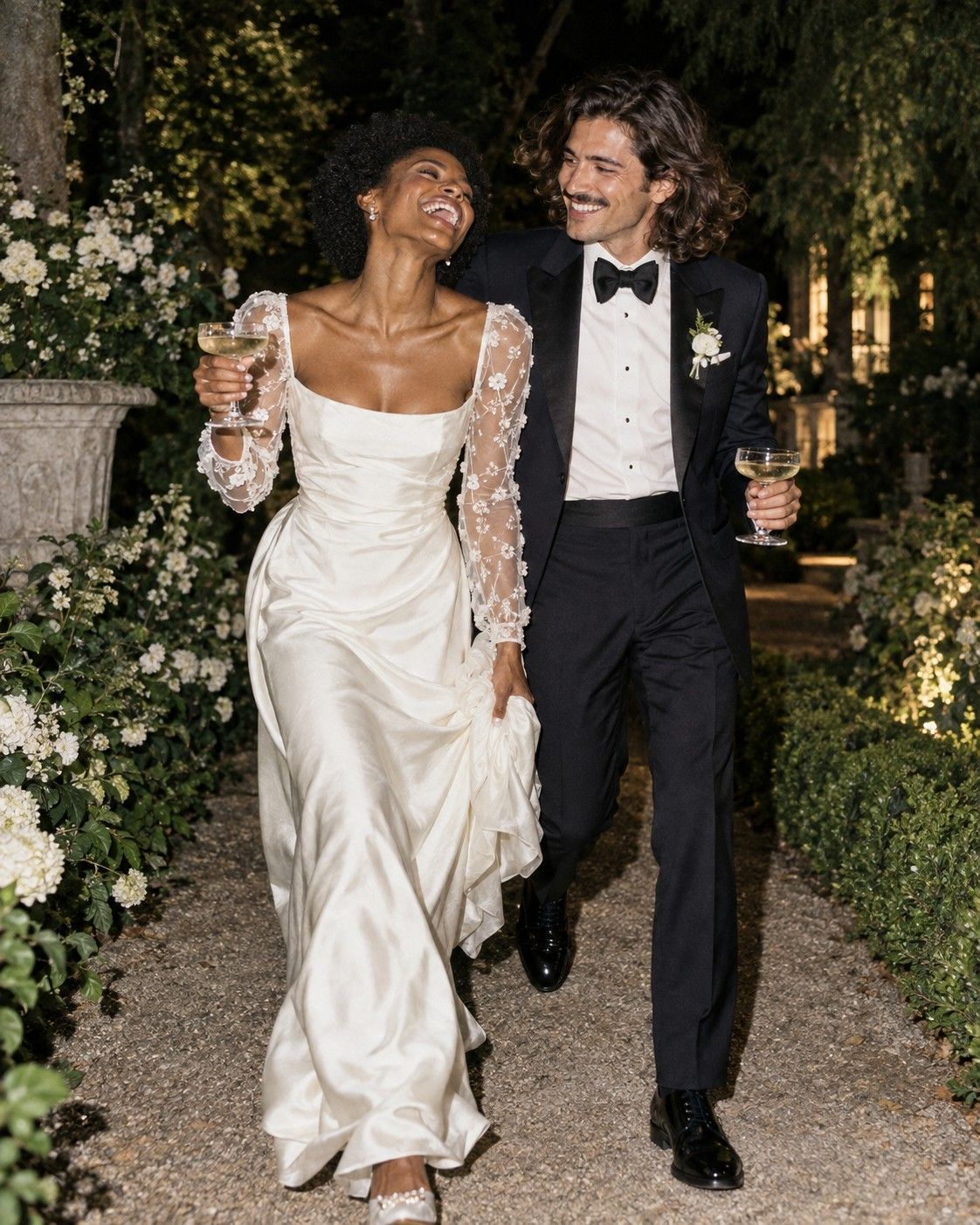 Una novia riendo y un novio que la mira sonriendo mientras caminan de noche por un sendero de jardín, con copas de champaña.